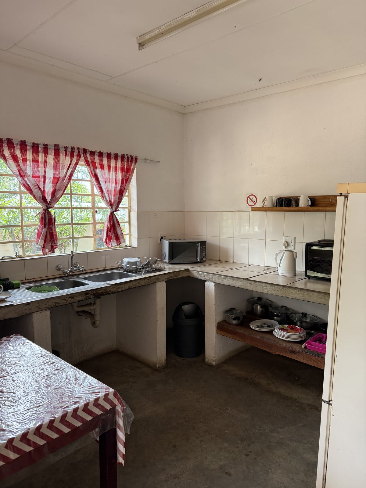 The self-catering kitchenette in a Kanaan lodge unit — tiled counters, two-plate hob, microwave, kettle, fridge and a small dining table beside a red-checked curtained window.