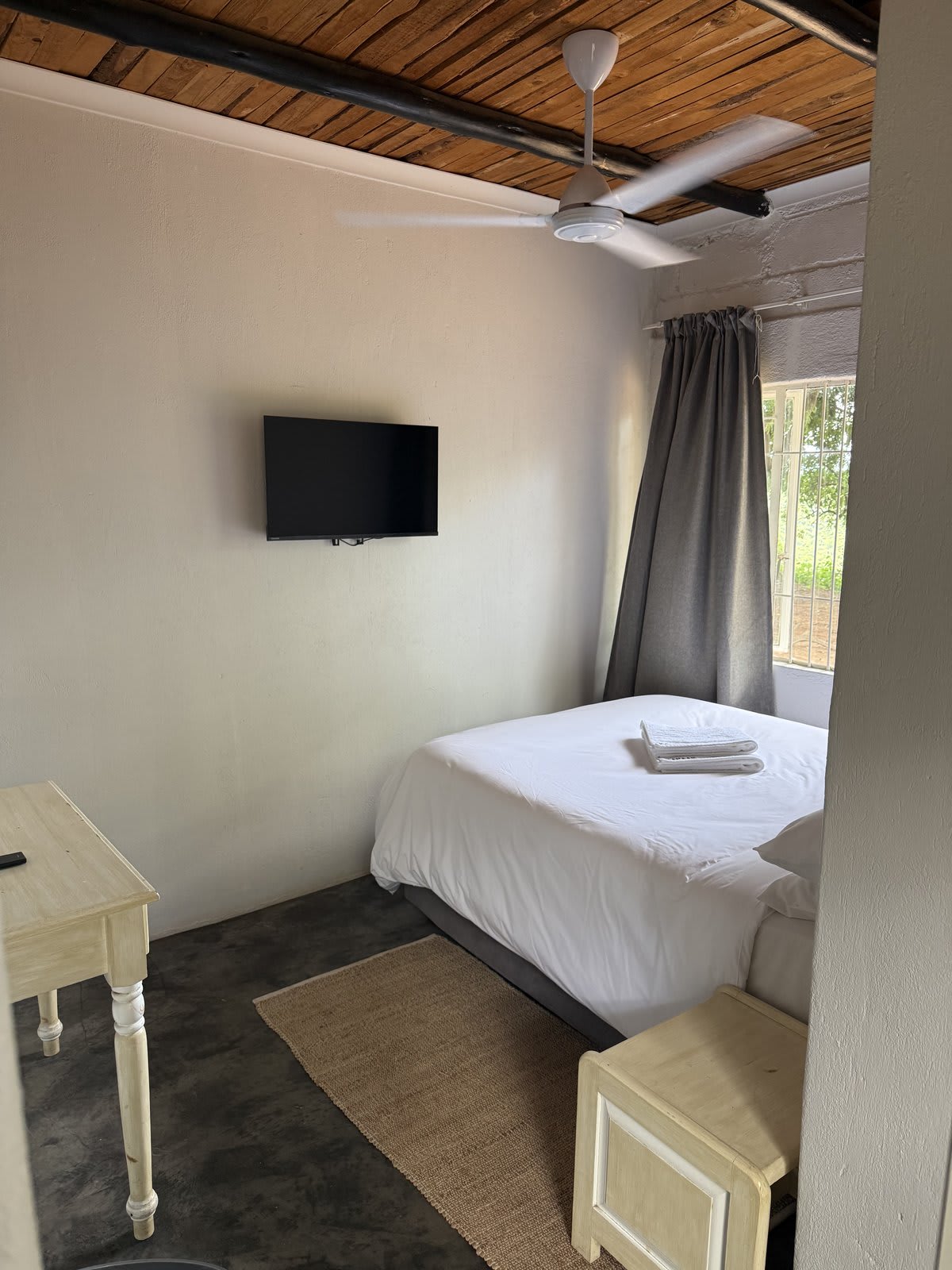 A double room in a renovated lodge unit at Kanaan — crisp white linen, wooden ceiling with exposed beams, ceiling fan and a wall-mounted TV.